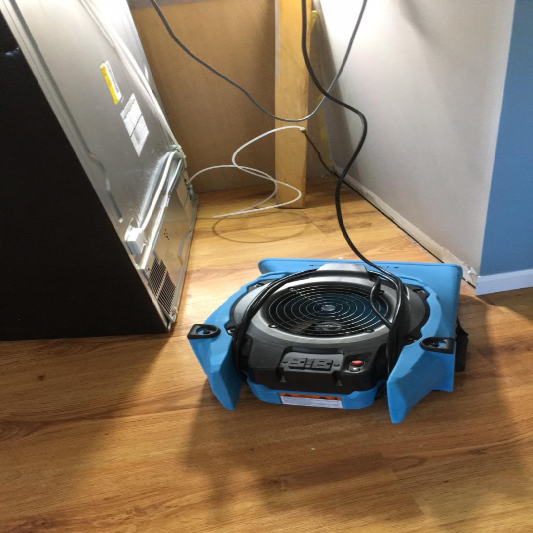 Air mover drying a hardwood floor behind a refrigerator after an ice maker line leak in New Jersey
